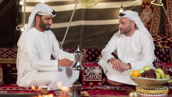 عادات وتقاليد أصيلة في تقديم المشروبات, السير على نهج الأجداد في إكرام الضيف,  رجل عربي خليجي اماراتي يرتدي الكندوره والغتره يقوم بصب القهوة من الدلة النحاسية لصديقه, توارث حسن الاستقبال والكرم, تقديم المنتجات المحلية للضيوف،المجلس الشعبي في الامارات