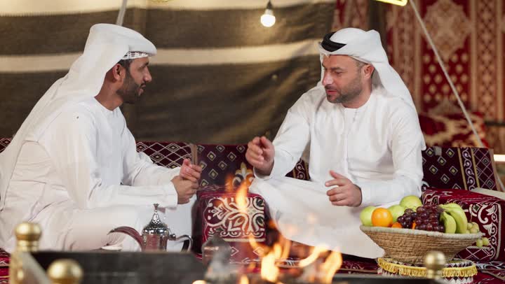 Good reception for friends in the People's Council, a young Arab Gulf Emirati man wearing a kandura and ghutra talking with his friend about daily topics, hand gestures indicating pointing and waving, honoring the guest and good hospitality and reception, chatting and talking about something