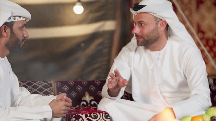الاستقبال الجيد للاصدقاء في المجلس الشعبي، شاب عربي خليجي اماراتي يرتدي الكندوره والغتره يقوم بالتحدث مع صديقه عن السوالف اليوميه، ايماءات يدين تدل على الاشاره والتلويح، اكرام الضيف و حسن الضيافه والاستقبال، التسامر والتحدث في امر ما