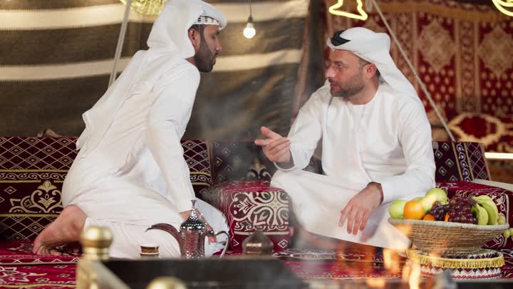 الاستقبال الجيد للاصدقاء في المجلس الشعبي، شاب عربي خليجي اماراتي يرتدي الكندوره والغتره يقوم بالتحدث مع صديقه عن السوالف اليوميه، ايماءات يدين تدل على الاشاره والتلويح، اكرام الضيف و حسن الضيافه والاستقبال، التسامر والتحدث في امر ما