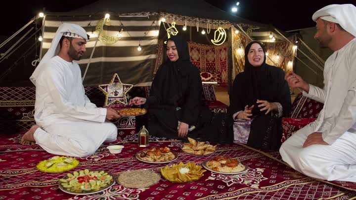 تبادل المأكولات والمقبلات بين الاصدقاء، التجمع حول مائده الافطار في شهر رمضان، تصوير من الاعلي لاصدقاء اماراتيون يجلسون في خيمه بدويه شعبيه يقومون بتقديم المقبلات والحلوي، المجلس الشعبي التقليدي، مأكولات واصناف متنوعه، التخيم الليلي في الصحراء