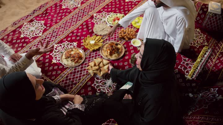 Gathering around the Iftar table during Ramadan, a top-down view of Emirati friends sitting in a traditional Bedouin tent exchanging food, the traditional Emirati Majlis, a variety of foods and appetizers, and a night camp in the desert.