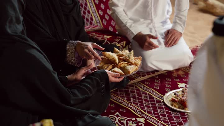A group of friends are having some appetizers, an Emirati man wearing a kandura and ghutra sitting with his friends, an Emirati woman is serving a plate full of appetizers to her friend, chatting and talking in groups, the principles of the traditional Majlis, honoring the guest and good reception
