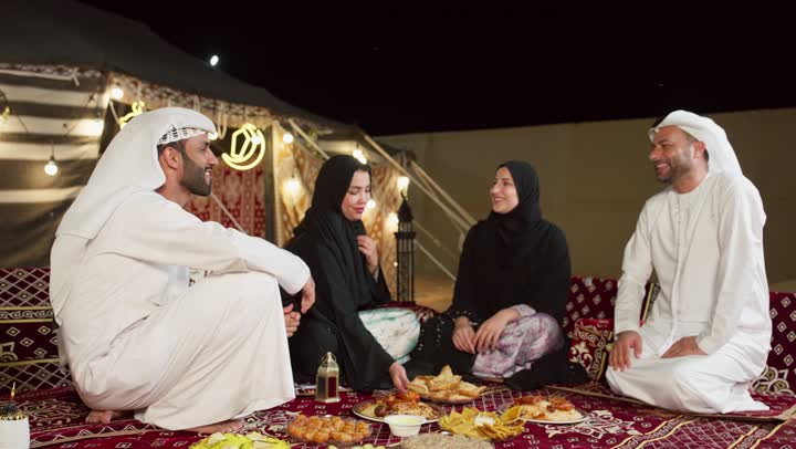 عائله اماراتيه في الصحراء، مائدة طعام كبيرة مليئة بأصناف المأكولات، مخيمات سياحية في المناطق البرية، خيمة مزينه بديكورات شهر رمضان الكريم، التخييم في المناطق الصحراويه في الامارات، امرأه تقوم بتقديم المقبلات لصديقتها، جلسة و كشتة برشبابية