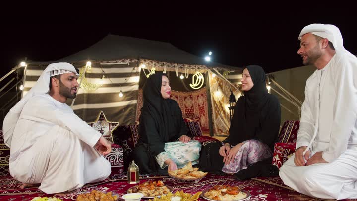 عائله اماراتيه في الصحراء، مائدة طعام كبيرة مليئة بأصناف المأكولات، مخيمات سياحية في المناطق البرية، خيمة مزينه بديكورات شهر رمضان الكريم، التخييم في المناطق الصحراويه في الامارات، امرأه تقوم بتقديم المقبلات لصديقتها، جلسة و كشتة برشبابية