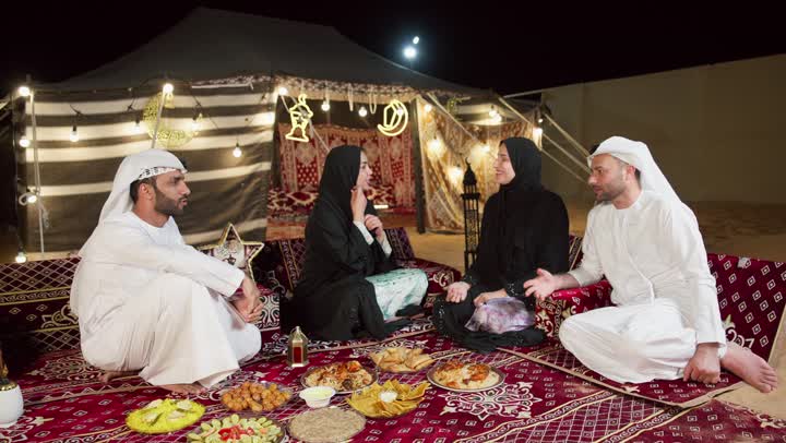 اصدقاء في المجلس الشعبي، شاب عربي خليجي اماراتي يرتدي الكندوره والغتره يقوم بالتسامر والتحدث في امر ما مع صديقه، افطار جماعي مع الاصدقاء، اكرام الضيف و حسن والاستقبال، صديقتان اماراتيتان تجلسان برفقه الاصدقاء