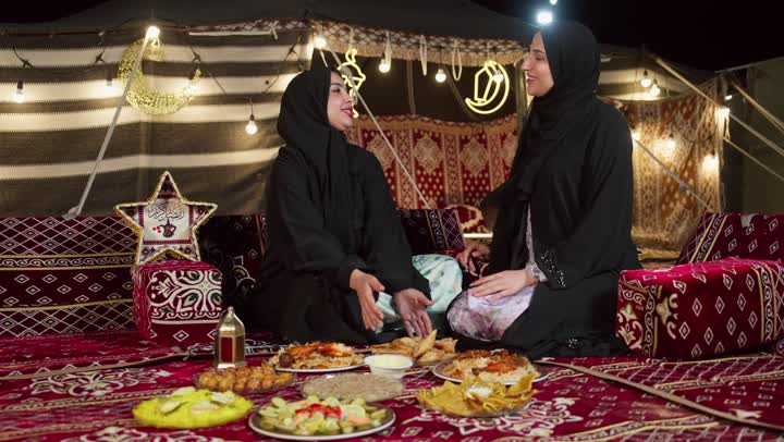 صديقتان سعوديتان ترتديان العباءه تجلسان في الخيمه ليلا، امرأه اماراتيه تقوم بتقديم المقبلات لصديقتها، مائدة طعام كبيرة مليئة بأصناف المأكولات، ديكورات شهر رمضان الكريم، التخييم في المناطق الصحراويه في الامارات