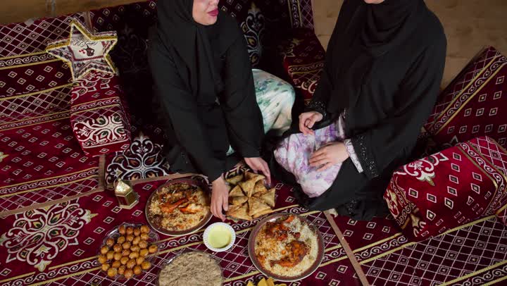 امرأه اماراتيه تقوم بتقديم المقبلات لصديقتها، مائدة طعام كبيرة مليئة بأصناف المأكولات، ديكورات شهر رمضان الكريم، التخييم في المناطق الصحراويه في الامارات، صديقتان سعوديتان ترتديان العباءه تجلسان في الخيمه ليلا، اصول وعادات المجلس الشعبي