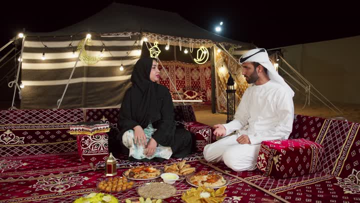 رجل اماراتي يرتدي الكندوره والغتره البيضاء يقدم لزوجته طبق من المقبلات، مائدة طعام كبيرة مليئة بأصناف المأكولات، مخيمات سياحية في المناطق البرية، خيمة مزينه بديكورات شهر رمضان الكريم، التخييم في المناطق الصحراويه في الامارات، جلسة و كشتة برشبابية