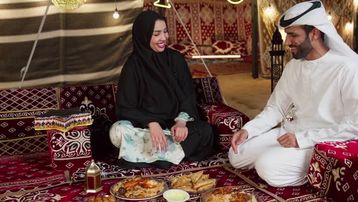 رجل اماراتي يرتدي الكندوره والغتره البيضاء يقدم لزوجته طبق من المقبلات، مائدة طعام كبيرة مليئة بأصناف المأكولات، مخيمات سياحية في المناطق البرية، خيمة مزينه بديكورات شهر رمضان الكريم، التخييم في المناطق الصحراويه في الامارات، جلسة و كشتة برشبابية