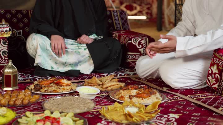 سيده اماراتيه تجلس برفقه زوجها امام مائده مليئه بالمأكولات المتنوعه، اطباق الارز الشعبي وصدور الدجاج المشوي، اصول وعادات المجلس الشعبي، خيمه عربيه تقليديه، رجل اماراتي مبتسم يرتدي الكندوره والغتره البيضاء يقوم بتقديم المقبلات لزوجته، التخييم الليلي