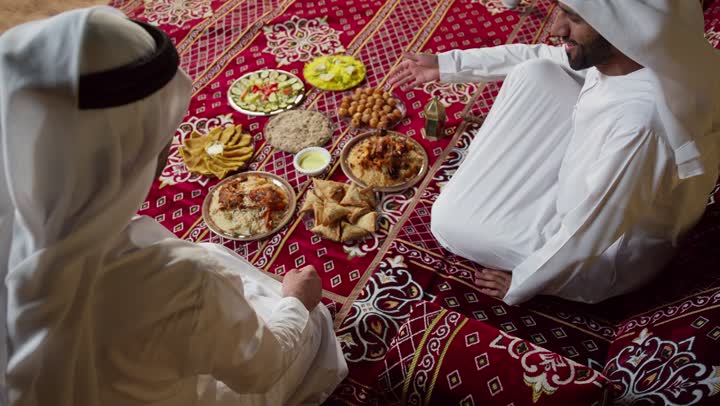رجلان عربيان خليجيان اماراتيان مبتسمان يرتديان الكندوره والغتره البيضاء يجلسان في خيمه مليئه بديكورات شهر رمضان، الكرم وحسن الضيافه والاستقبال، شاب اماراتي يقدم طبق من الحلوي لصديقه، التخييم الليلي في اجواء رمضانيه، اصول وعادات المجلس الشعبي