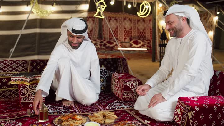 رجلان عربيان خليجيان اماراتيان مبتسمان يرتديان الكندوره والغتره البيضاء يجلسان في خيمه مليئه بديكورات شهر رمضان، الكرم وحسن الضيافه والاستقبال، شاب اماراتي يقدم طبق من الحلوي لصديقه، التخييم الليلي في اجواء رمضانيه، اصول وعادات المجلس الشعبي