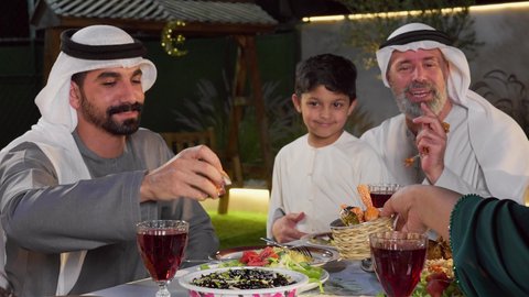قضاء أوقات ممتعة مع العائلة في الهواء الطلق، مائدة إفطار مليئة بالأصناف والمأكولات الشهية، عائلة عربية خليجية إماراتية تقوم بتناول الافطار السعادة و السرور، الشهر الكريم والأجواء الرمضانية الروحانية، رجل عربي خليجي يقوم بتناول قطع من الحلوي