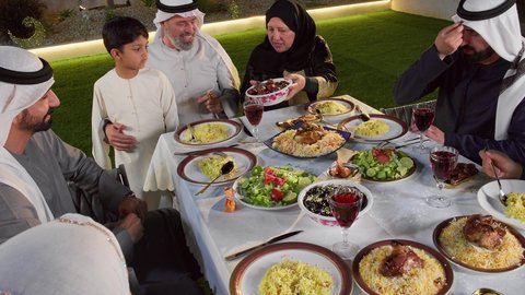 تجمع العائلة على سفرة الاكل, صورة مقربة لامرأة تقوم بتقديم طبق من التمر, ايماءات السعادة و السرور بقدوم شهر رمضان المبارك, الاحتفال بالاعياد و المناسبات الاسلامية, عائلة عربية خليجية اماراتية تجلس على الطاولة المليئة بالاطباق الشهية