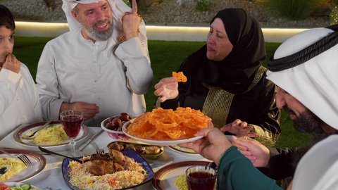 نقاش وحوار عائلي، حسن الضيافة و الاكرام، مائده طعام مليئه بأصناف المأكولات و المقبلات الشهية، ديكور وأجواء رمضانية، تجمع أسرة عربية خليجية اماراتية حول سفرة الإفطار في شهر رمضان المبارك، ايماءات السعادة والسرور، تصويرمقرب لامراة خليجيه تقوم بتقديم طبق من الحلويات الشرقية