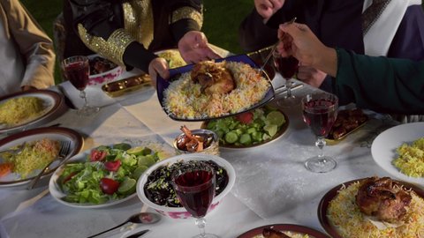 تجمع العائلة على سفرة الاكل, صورة مقربة لامرأة تقوم بسكب الطعام في الصحون و توزيعه على افراد الاسرة, ايماءات السعادة و السرور بقدوم شهر رمضان المبارك, الاحتفال بالاعياد و المناسبات الاسلامية, عائلة عربية خليجية اماراتية تجلس على الطاولة المليئة بالاطباق الشهية