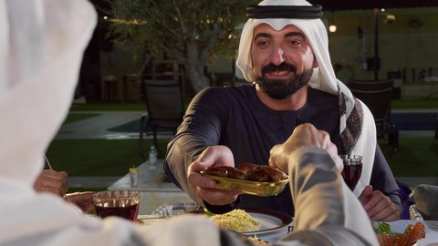 أجواء دينية رمضانية، مشاركة الأطعمة و إكرام الضيف، أداء فريضة الصيام، العزائم والولائم في الأعياد والمناسبات الإسلامية، تصوير مقرب لرجل عربي خليجي اماراتي يرتدي الزي التقليدي يقوم بتقديم طبق من التمر، ايماءات السعادة و السرور