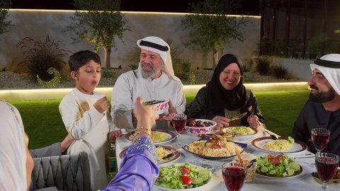 مائده طعام مليئه بأصناف المأكولات و المقبلات الشهية،رجل عربي خليجي اماراتي يرتدي الزي التقليدي يقوم بتقديم طبق من ورق العنب، تجمع أسرة عربية خليجية اماراتية حول سفرة الإفطار في شهر رمضان المبارك، ايماءات السعادة والسرور، حسن الضيافة و الاكرام