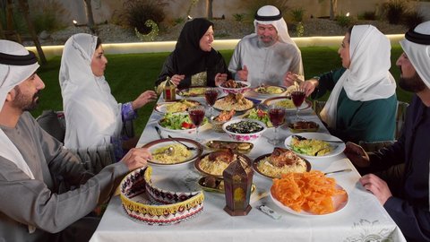 تجمع العائلة على سفرة الاكل, تصوير لامرأة تقوم بتقديم التمر لافراد الاسرة, ايماءات السعادة و السرور بقدوم شهر رمضان المبارك, الاحتفال بالاعياد و المناسبات الاسلامية, عائلة عربية خليجية اماراتية تجلس على الطاولة المليئة بالاطباق الشهية