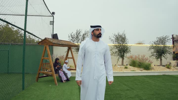 النظر الي شئ ما، رجل اماراتي يرتدي الكندوره والغتره البيضاء يسير في حديقه المنزل، المشي باستقامه وثقه في النفس، الاهتمام بالمظهر الخارجي، ام عربيه خليجيه تقوم بحمل الهاتف في الحديقه برفقه ابنها، الجلوس فى حديقه المنزل، استخدام التقنيات الحديثه