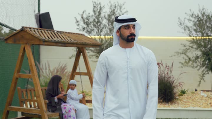 النظر الي شئ ما، رجل اماراتي يرتدي الكندوره والغتره البيضاء يسير في حديقه المنزل، المشي باستقامه وثقه في النفس، الاهتمام بالمظهر الخارجي، ام عربيه خليجيه تقوم بحمل الهاتف في الحديقه برفقه ابنها، الجلوس فى حديقه المنزل، استخدام التقنيات الحديثه