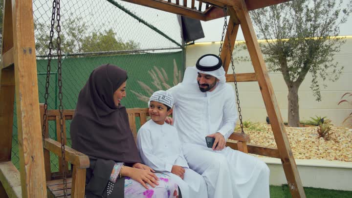 رجل اماراتي يرتدي الكندوره والغتره البيضاء يجري مكالمه هاتفيه، قضاء أوقات مليئه بالحب والموده، ام عربيه خليجيه تقوم بالمداعبه واللعب في الحديقه برفقه ابنها، الجلوس فى حديقه المنزل، استخدام التقنيات الحديثه