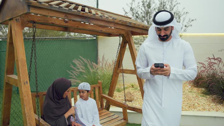 Emirati man wearing white kandura and ghutra using mobile phone, spending time full of love and affection, Arab Gulf mother talking and sitting in the garden with her son, sitting in the home garden, using modern technology
