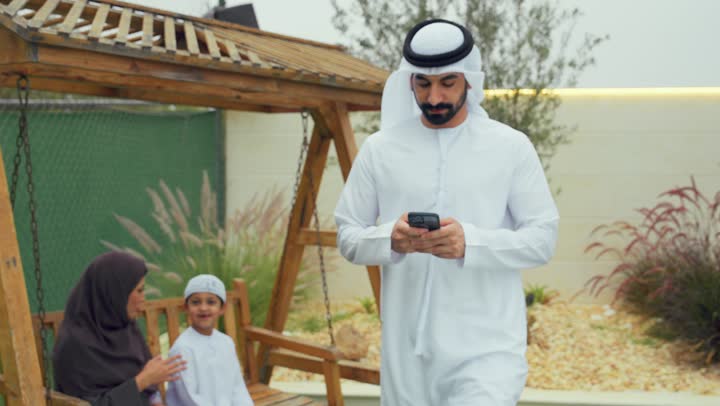 رجل اماراتي يرتدي الكندوره والغتره البيضاء يقوم باستخدام الهاتف المحمول، قضاء أوقات مليئه بالحب والموده،ام عربيه خليجيه تقوم باالتحدث والجلوس في الحديقه برفقه ابنها، الجلوس فى حديقه المنزل، استخدام التقنيات الحديثه