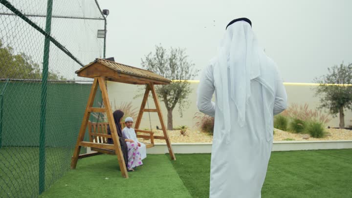رجل اماراتي برفقه عائلته يقوم بالتقاط صوره لزوجته وابنه عبر الهاتف المحمول، قضاء أوقات مليئه بالحب والموده، زوجان عربيان خليجيان اماراتيان يجلسان برفقه ابنهم، الجلوس فى حديقه المنزل، استخدام التقنيات الحديثه، ايماءه الاشاره الي الهاتف