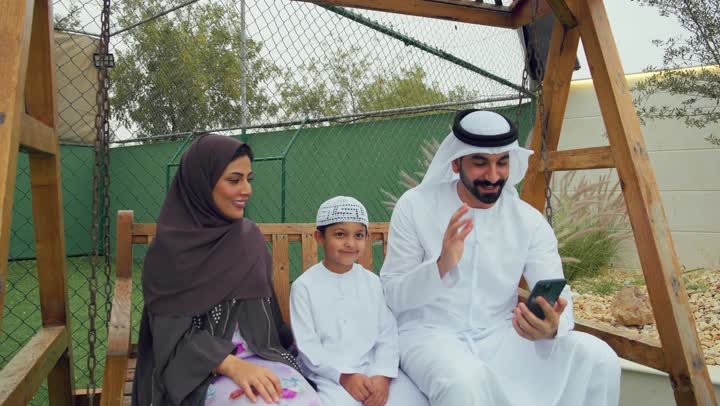 قضاء أوقات مليئه بالحب والموده، زوجان عربيان خليجيان اماراتيان يجلسان برفقه ابنهم، الجلوس فى حديقه المنزل، رجل اماراتي برفقه عائلته يقوم بمكالمه فيديوعبر الهاتف المحمول، التواصل عن بعد، استخدام التقنيات الحديثه