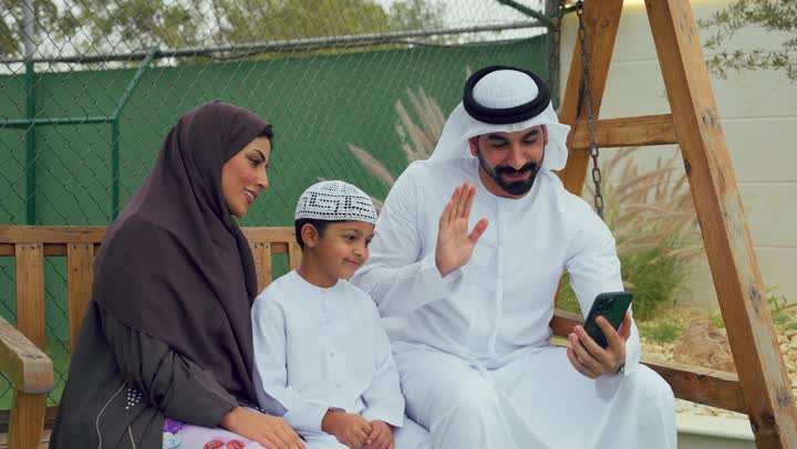 قضاء أوقات مليئه بالحب والموده، زوجان عربيان خليجيان اماراتيان يجلسان برفقه ابنهم، الجلوس فى حديقه المنزل، رجل اماراتي برفقه عائلته يقوم بمكالمه فيديوعبر الهاتف المحمول، التواصل عن بعد، استخدام التقنيات الحديثه