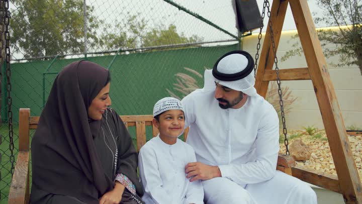 استخدام التقنيات الحديثه، زوجان عربيان خليجيان اماراتيان يجلسان برفقه ابنهم ويقومان بتوثيق اللحظات السعيده، الجلوس فى حديقه المنزل، امرأه اماراتيه برفقه عائلتها تقوم بالتقاط صور تذكاريه عبر الهاتف المحمول