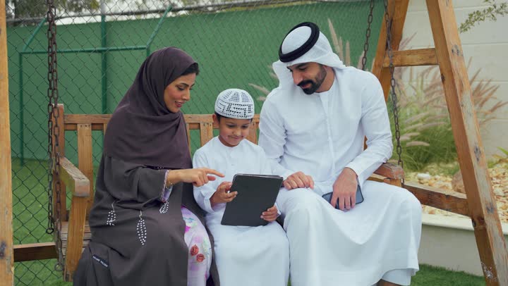 زوجان عربيان خليجيان اماراتيان يجلسان برفقه ابنهم ويقومان بتعليمه علي التابلت، استخدام التقنيات الحديثه، دمج التكنولوجيا مع التعليم عن بعد، الجلوس فى حديقه المنزل، ولد اماراتي يستخدم الجهاز اللوحي برفقه والديه