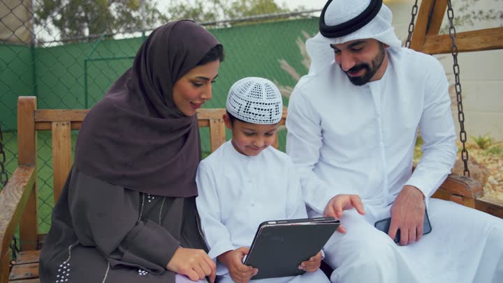 زوجان عربيان خليجيان اماراتيان يجلسان برفقه ابنهم ويقومان بتعليمه علي التابلت، استخدام التقنيات الحديثه، دمج التكنولوجيا مع التعليم عن بعد، الجلوس فى حديقه المنزل، ولد اماراتي يستخدم الجهاز اللوحي برفقه والديه