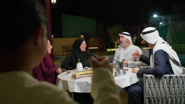 طفل عربي خليجي اماراتي يمسك بيده فانوس معدني ملون يقدمه لجده، استخدام الديكورات الفنية في الزينة، جلسة أسرية في حديقة المنزل، التمتع بالهواء الطلق، عائله اماراتيه تجلس في احد الاماكن الخارجيه، التسامر مع افراد العائله