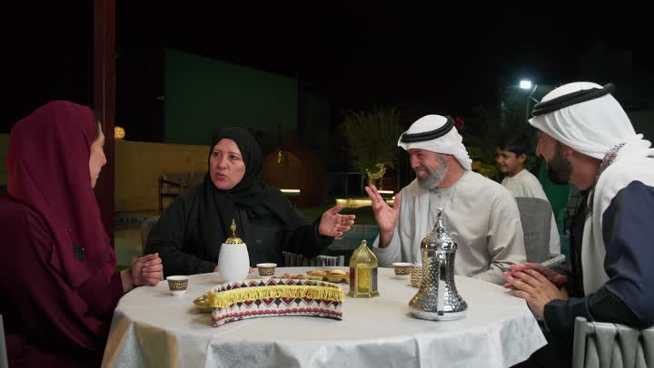 فطور رمضاني عائلي في مطعم مزين بديكورات رمضانية, اسلاميات وعبادة,عائلة عربية خليجية إماراتية تجلس على سفرة الطعام تقوم بالتحدث والتسامر في اجواء شهر رمضان الكريم, أسرة اماراتيه مستمتعه بالجلوس في الخارج، ولد اماراتي مبتسم يحمل فانوس معدني مزخرف ولون