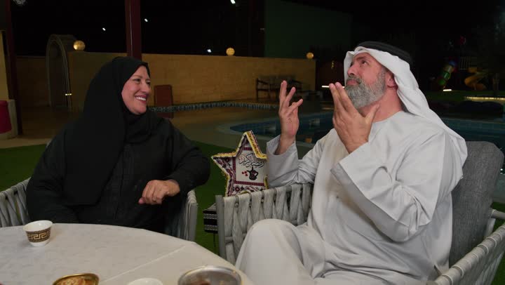 An elderly Emirati man and his wife praying and making wishes, a happy Ramadan religious family atmosphere, colorful decorative metal lanterns on the dining table, Islamic event decorations, performing the fasting duty in the holy month of Ramadan, an Emirati Gulf Arab family gathered around the dining table