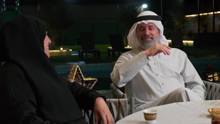 رجل مسن اماراتي برفقه زوجته يتبادلان الحديث، أجواء عائلية دينية رمضانية سعيدة، فوانيس معدنية مزخرفة ملونة على سفرة الطعام، زينة المناسبات الإسلامية، أداء فريضة الصيام في شهر رمضان المبارك، عائلة عربية خليجية اماراتيه مجتمعة حول مائدة الطعام
