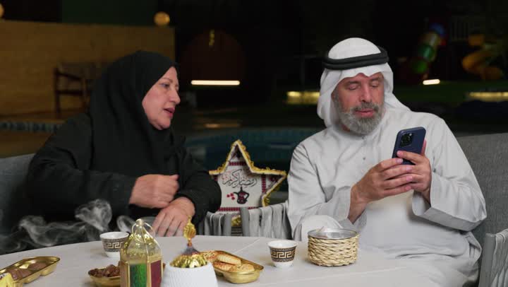 زوجان عربيان خليجيان اماراتيان على مائدة الافطار في شهر رمضان، ايماءات يدين تدل علي التمني والدعاء، جلسة عائلية رمضانية روحانية، زينة وديكور شهر رمضان المبارك، استخدام تطبيقات الهاتف لطلب المأكولات والمشروبات، اشتخدام الاجهزه والتقنيات الحديثه