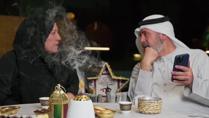 استخدام خشب العود في المناسبات والأعياد، منظر الدخان المنتشر من البخور، زوجان عربيان اماراتيان يجلسان علي طاوله الافطارالرمضاني، رجل مسن برفقه زوجته يستخدم الهاتف المحمول، استخدام التقنيات الحديثه، افطارخارجي مع العائله