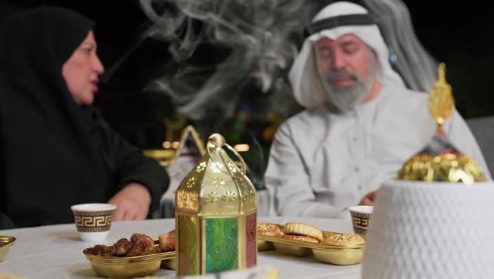 استخدام خشب العود في المناسبات والأعياد، منظر الدخان المنتشر من البخور، زوجان عربيان اماراتيان يجلسان علي طاوله الافطارالرمضاني، رجل مسن برفقه زوجته يستخدم الهاتف المحمول، استخدام التقنيات الحديثه، افطارخارجي مع العائله