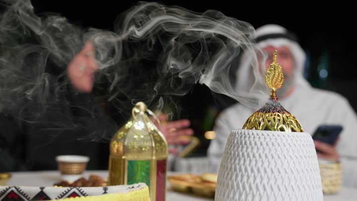 استخدام خشب العود في المناسبات والأعياد، منظر الدخان المنتشر من البخور، زوجان عربيان اماراتيان يجلسان علي طاوله الافطارالرمضاني، رجل مسن برفقه زوجته يستخدم الهاتف المحمول، استخدام التقنيات الحديثه، افطارخارجي مع العائله