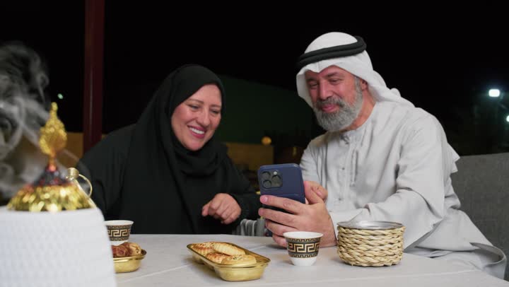 امرأه سعوديه ترتدي عباءه سوداء مسنه تتحدث مع زوجها, افطار رمضاني في الخارج, ديكورات وزينه شهر رمضان، التسامروقضاء اوقات في الخارج،رجل مسن عربي خليجي اماراتي يمسك الهاتف بايماءه الضحك والسعاده، استخدام الهاتف لاجراء مكالمه فيديو
