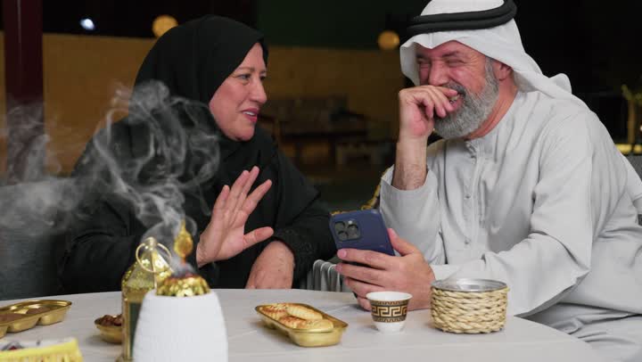 امرأه سعوديه ترتدي عباءه سوداء مسنه تتحدث مع زوجها, افطار رمضاني في الخارج, ديكورات وزينه شهر رمضان، التسامروقضاء اوقات في الخارج،رجل مسن عربي خليجي اماراتي يمسك الهاتف بايماءه الضحك والسعاده، استخدام الهاتف لاجراء مكالمه فيديو