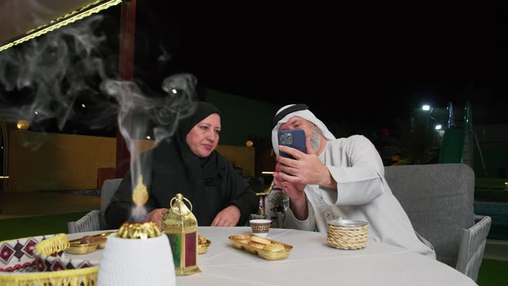 مفهوم المحبة والترابط الأسري، رجل عربي خليجي اماراتي يجلس برفقه زوجته على مائدة الطعام يقوم بمكالمه فيديوعبر الهاتف المحمول، اكواب المشروبات الساخنه، ديكورات شهر رمضان، استخدام التقنيات الحديثه