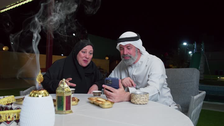 مفهوم المحبة والترابط الأسري، رجل عربي خليجي اماراتي يجلس برفقه زوجته على مائدة الطعام يقوم بمكالمه فيديوعبر الهاتف المحمول، اكواب المشروبات الساخنه، ديكورات شهر رمضان، استخدام التقنيات الحديثه
