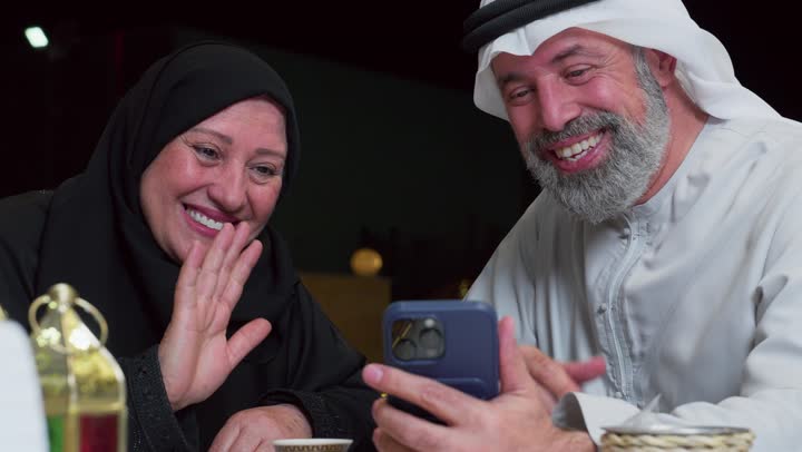 مفهوم المحبة والترابط الأسري، رجل عربي خليجي اماراتي يجلس برفقه زوجته على مائدة الطعام يقوم بمكالمه فيديوعبر الهاتف المحمول، اكواب المشروبات الساخنه، ديكورات شهر رمضان، استخدام التقنيات الحديثه