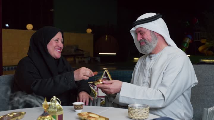 تناول التمر لكسر الصيام، رجل مسن عربي خليجي اماراتي يرتدي الكندوره والغتره البيضاء يقضي وقت ممتع مع زوجته على مائدة مليئة بالأطعمة والمشروبات الشهية, جلسة عائلية رمضانية روحانية، زينة وديكور شهر رمضان المبارك