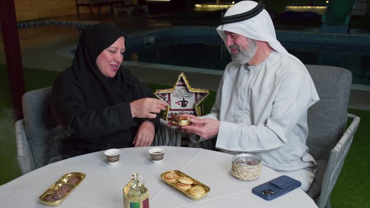 تناول التمر لكسر الصيام، رجل مسن عربي خليجي اماراتي يرتدي الكندوره والغتره البيضاء يقضي وقت ممتع مع زوجته على مائدة مليئة بالأطعمة والمشروبات الشهية, جلسة عائلية رمضانية روحانية، زينة وديكور شهر رمضان المبارك